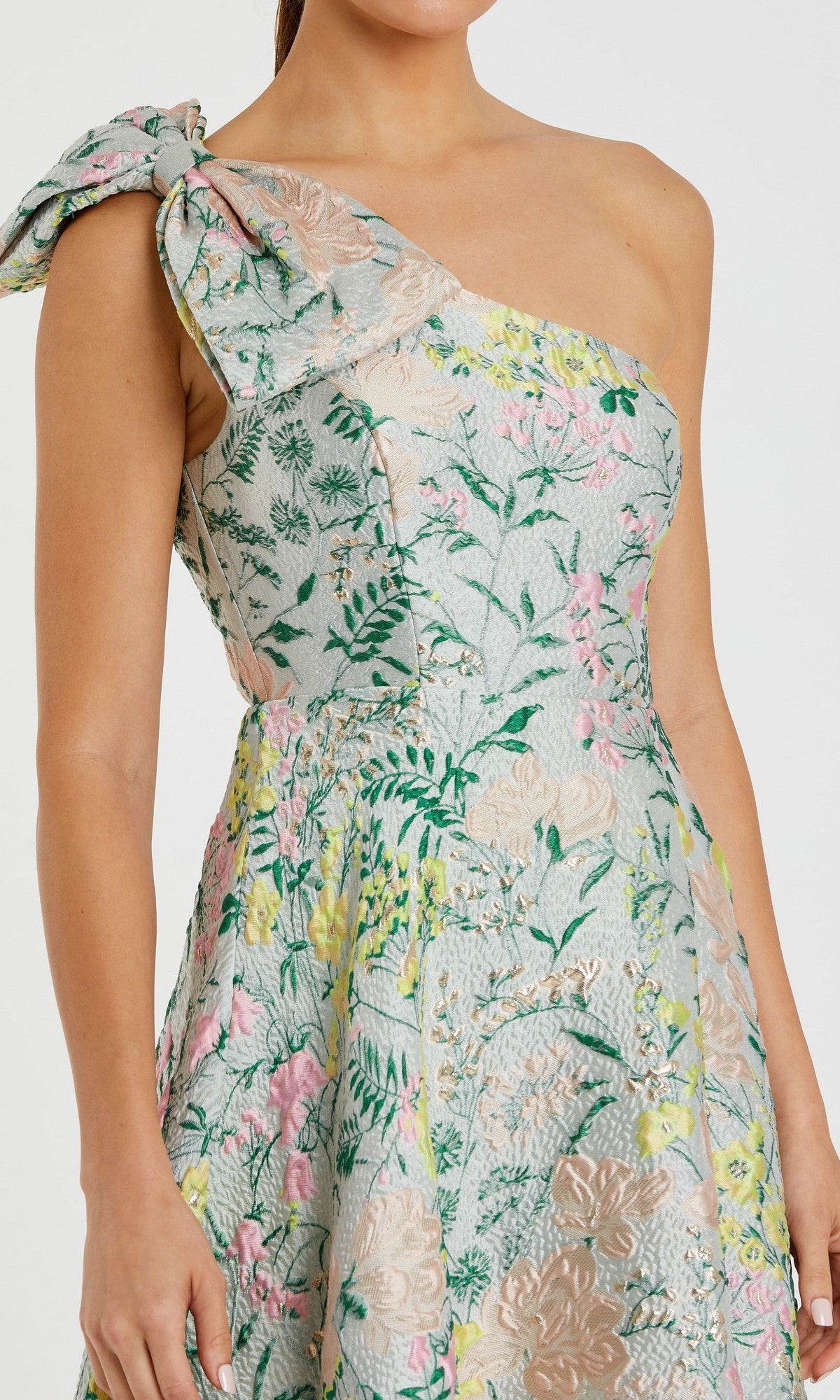 A woman wears the Mac Duggal 11919 long formal dress, featuring a one-shoulder design with a bow, pastel floral brocade fabric, and green leaves on a light background—an elegant choice for special occasions.